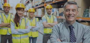 man in warehouse with people in hard hats smiling in background